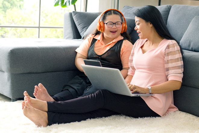 Two young women with dark hair, sitting together on living room floor, smiling as they look at a computer screen. One woman has a developmental disability and is wearing headphones. 